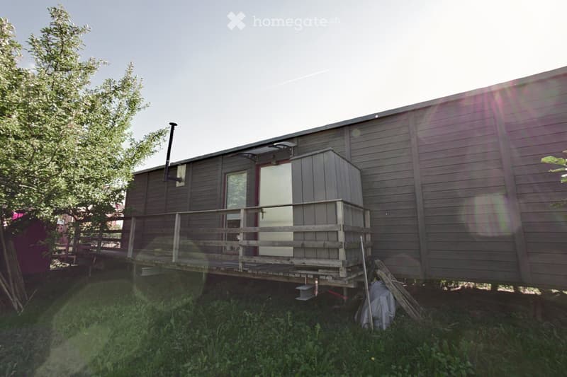 A small, single-story house with wooden siding, a raised wooden deck, and a small utility shed attached to the side.