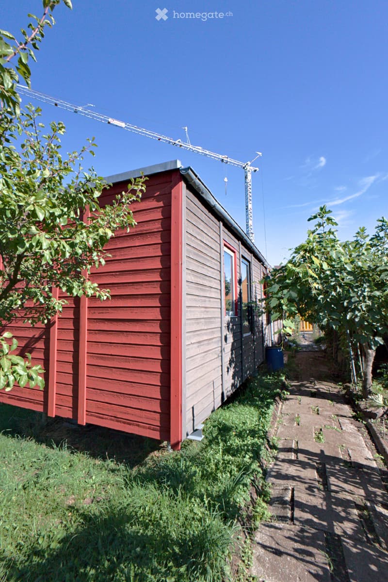 A small, red wooden building with a grey roof, surrounded by greenery and a paved pathway.
