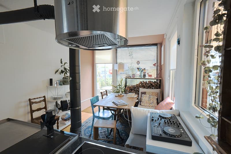 A cozy, well-lit living space with a sofa, DJ turntable, dining table, chairs, shelves, and a wood pile near the window.