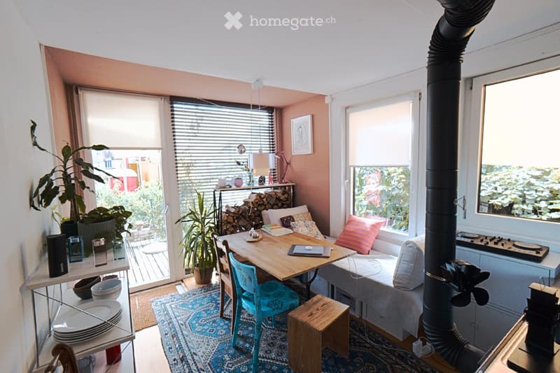 Cozy living room with a sofa, wooden table, chair, and a small wooden stove. The room features large windows with blinds, a bookshelf, and decorative plants.