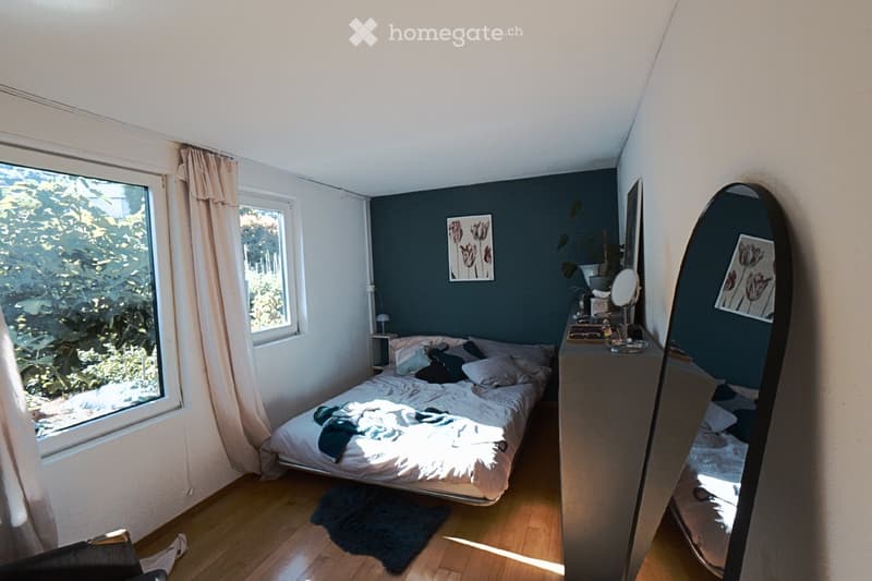 Cozy bedroom with a double bed, nightstand, mirror, and artwork on the walls. The room features wooden flooring and large windows with curtains, allowing natural light to fill the space.