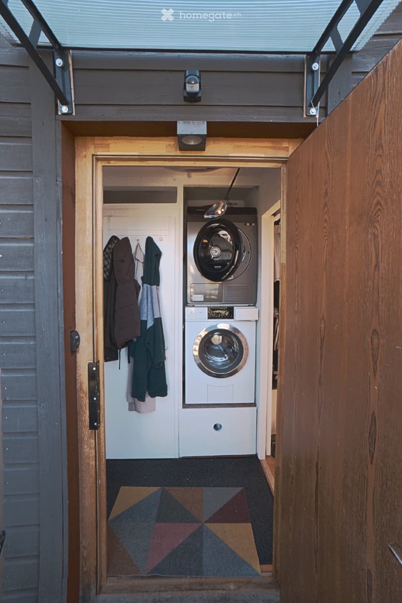 A narrow hallway with wooden walls and a skylight above, leading to a small laundry area with a washer and dryer, and a coat rack with jackets and coats.