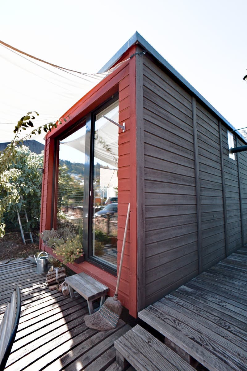 A small wooden shed with a red exterior and a large window, situated on a wooden deck. The deck features a broom, a watering can, a small stool, and a flower pot with plants. Wooden planks and logs are also visible on the deck.