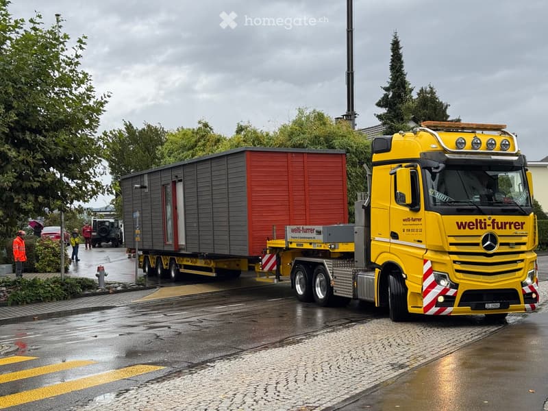 A large yellow truck with a trailer carrying a modular building structure, parked on a wet street with trees and a few people nearby.