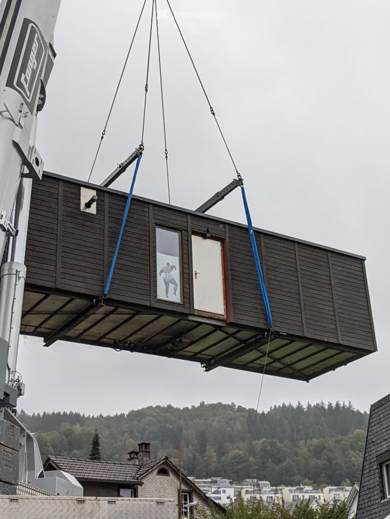A prefabricated wooden house being lifted by a crane, featuring a window and a door, with a forested hill in the background.