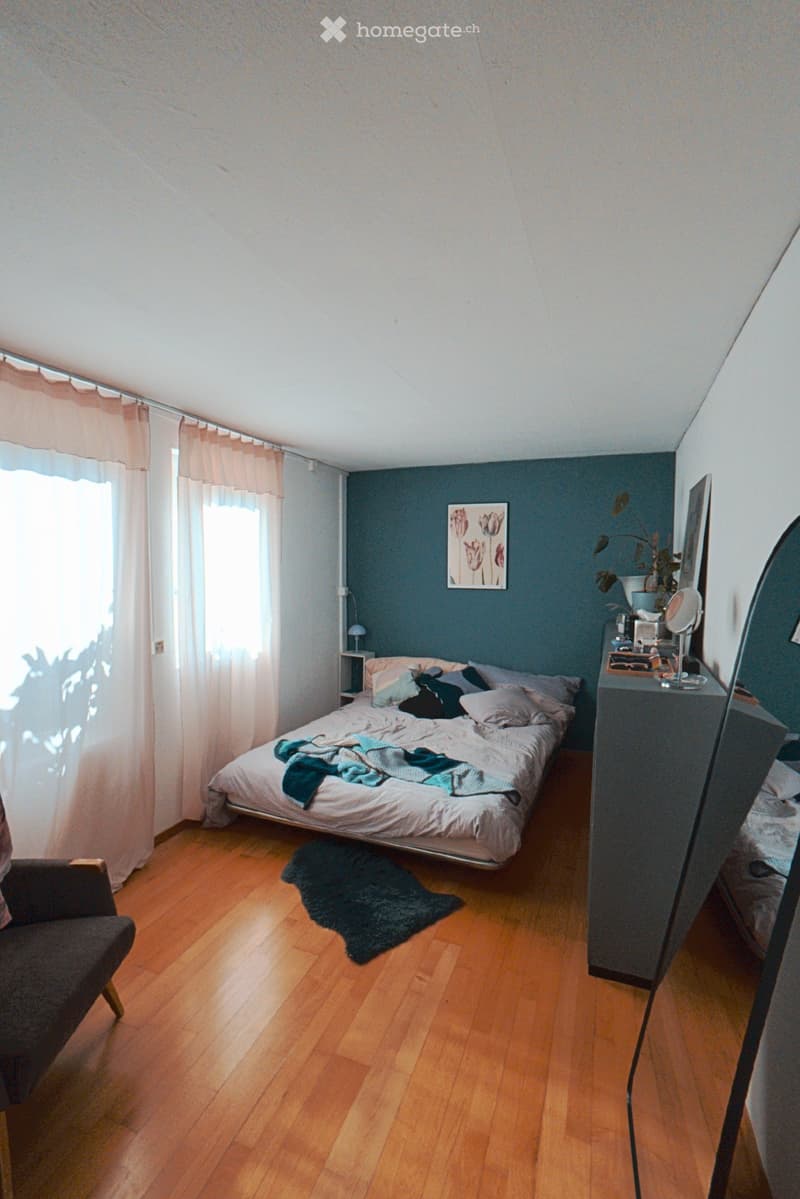 Cozy bedroom with wooden flooring, a large bed with pillows and blankets, a dark blue accent wall, a nightstand, a chair, and a mirror.