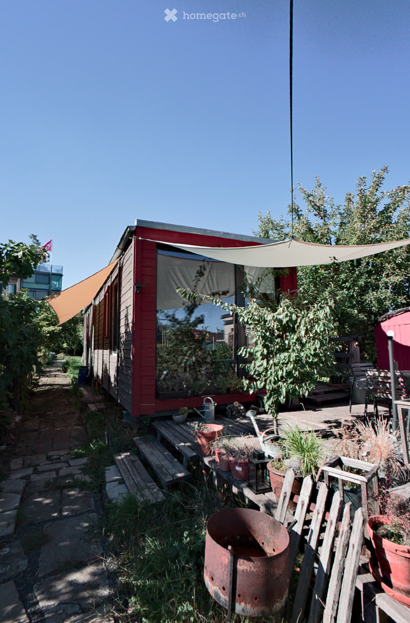 A small red house with a covered patio area, surrounded by greenery and potted plants. The patio includes wooden benches, a metal fire pit, and various garden tools.