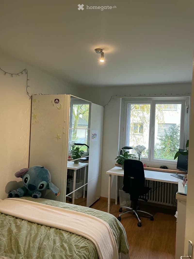 A small, well-lit bedroom with a bed, desk, and chair. The room features a large window with a view of greenery, a wardrobe, and a small plant on the desk.