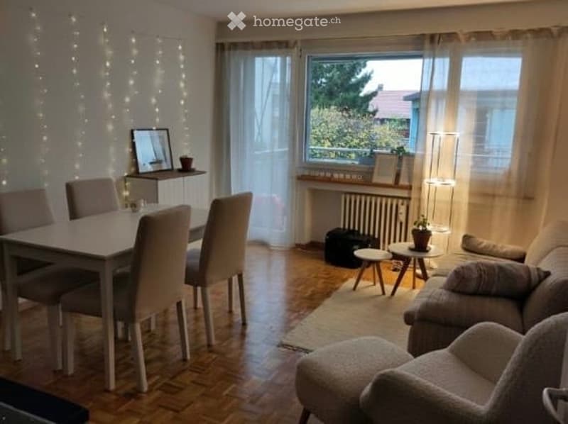 Bright, open-plan living and dining area with wooden flooring, large windows, a sofa, ottoman, dining table with chairs, and decorative items including a mirror and small plant.
