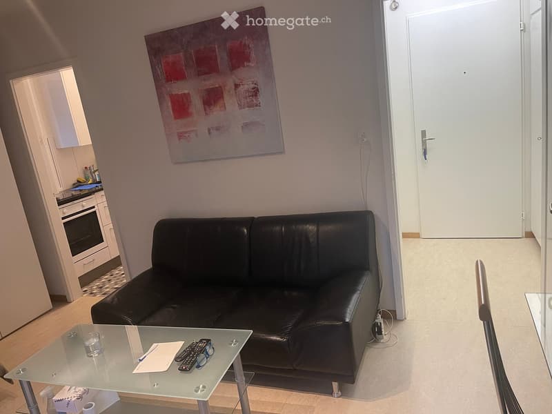 A modern living room with a black leather sofa, a glass coffee table, and a large abstract painting on the wall. A small kitchen area is visible in the background.