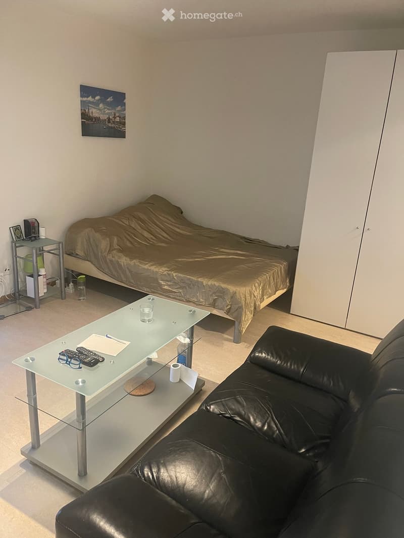 A furnished room with a bed, a sofa, a glass coffee table, and a small side table with a picture frame and other items.