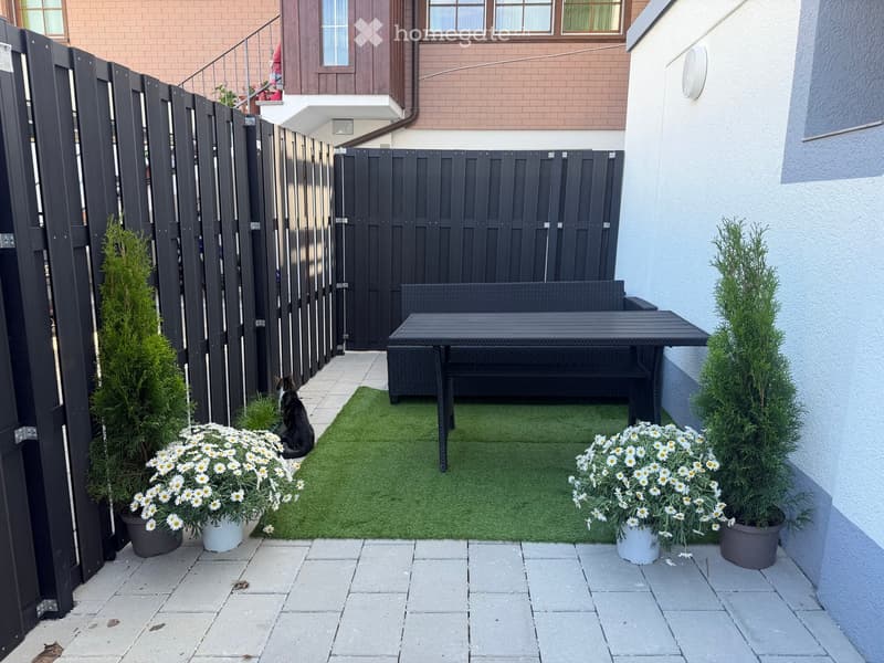 A small, enclosed patio with artificial grass, a black wicker sofa, a black table, potted flowers, and a cat sitting on the grass.