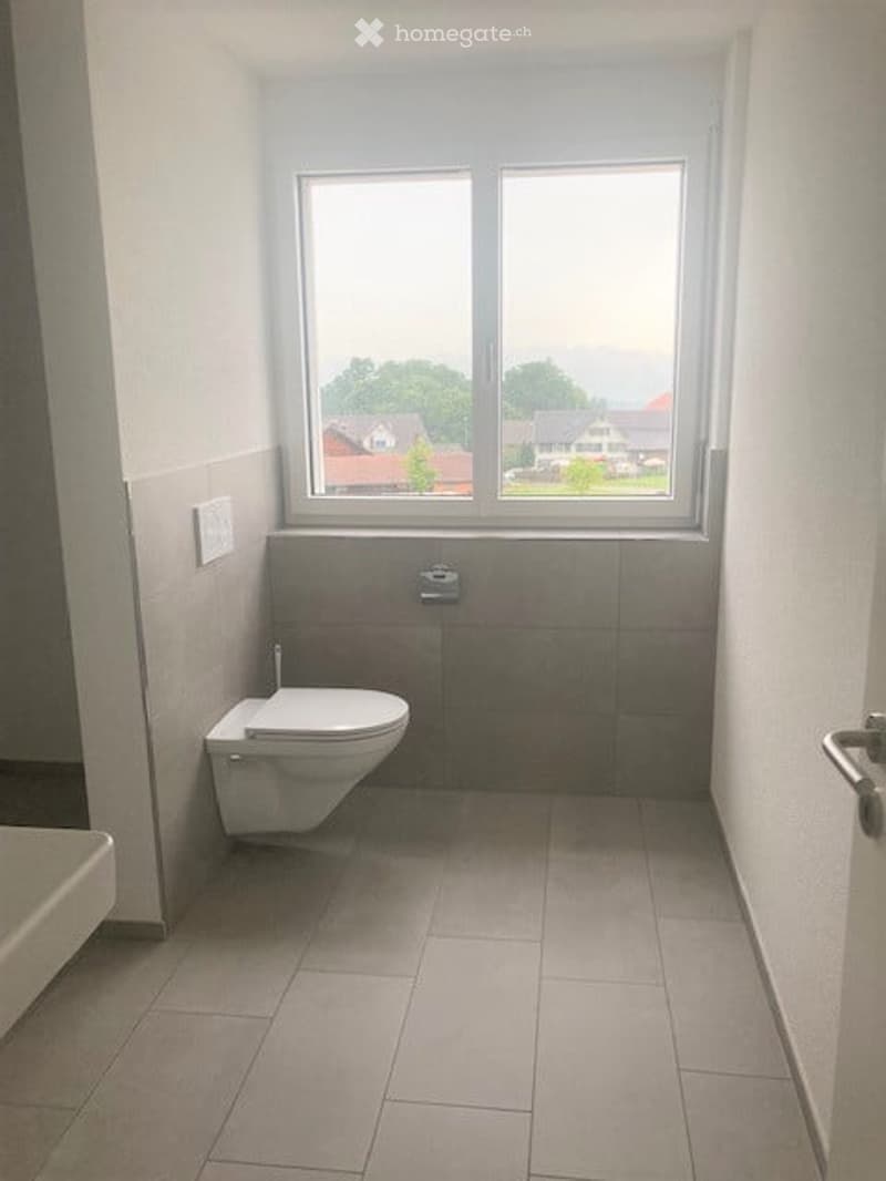 Modern bathroom with a toilet, large window, tiled walls and floor, and a light switch on the wall.