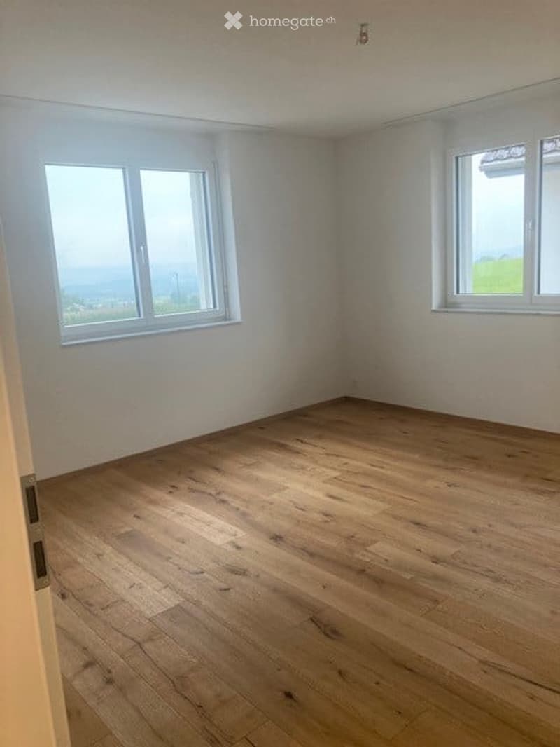 Empty room with wooden flooring and white walls, featuring two windows with white frames and a view of greenery outside.