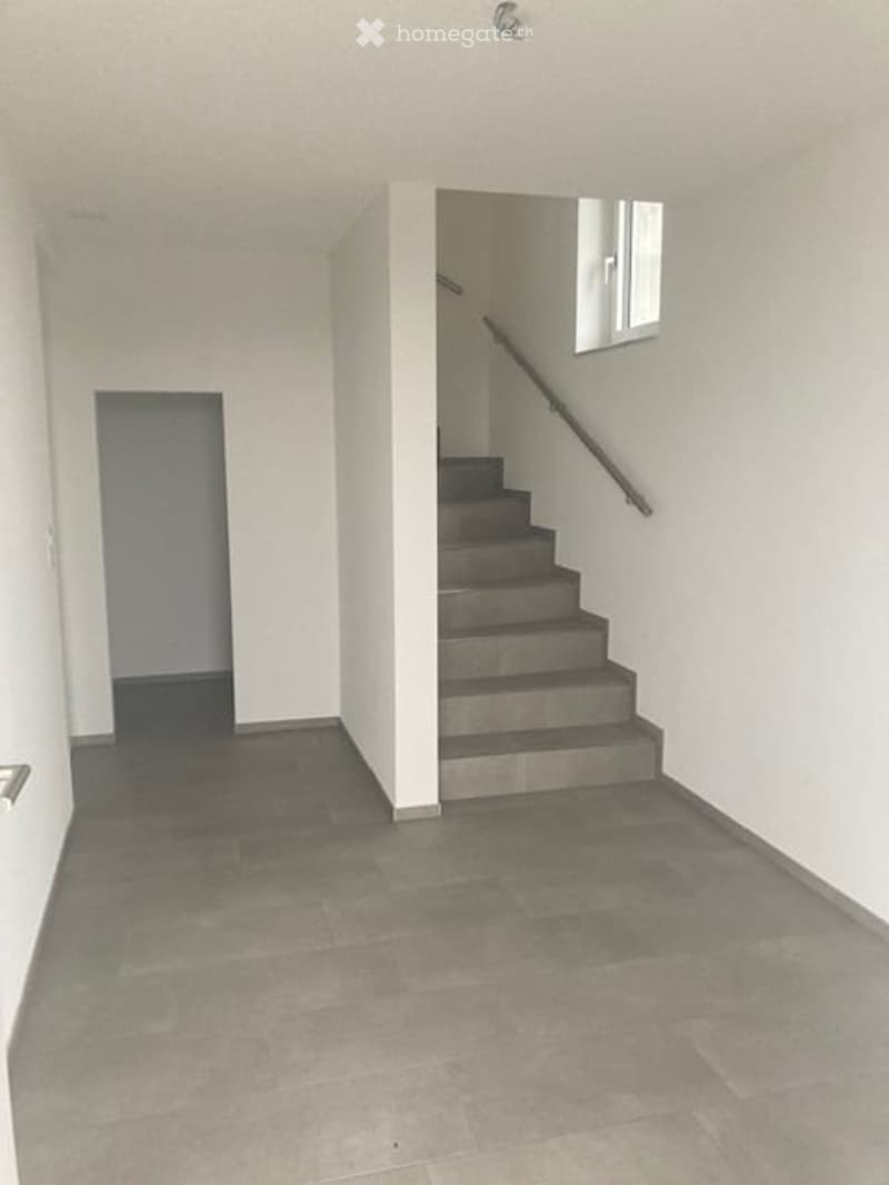 Empty hallway with grey tiled floor, white walls, and a staircase leading upwards with a metal railing.