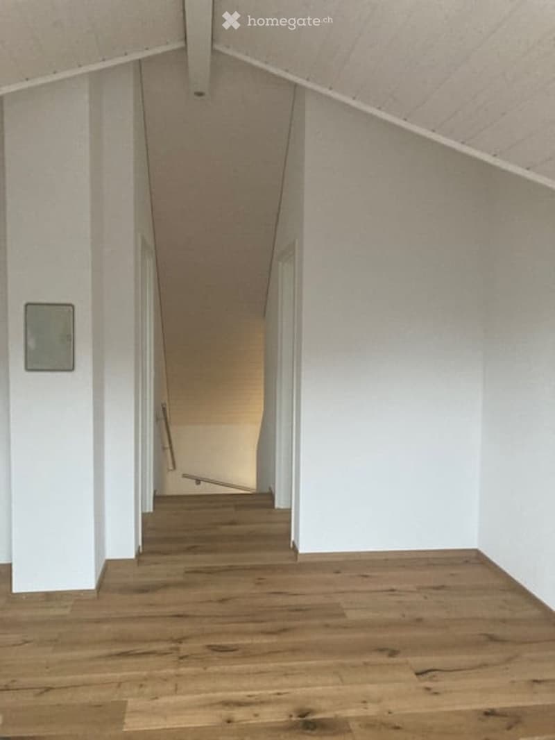 Empty hallway with wooden flooring and white walls, leading to another room with a slanted ceiling.