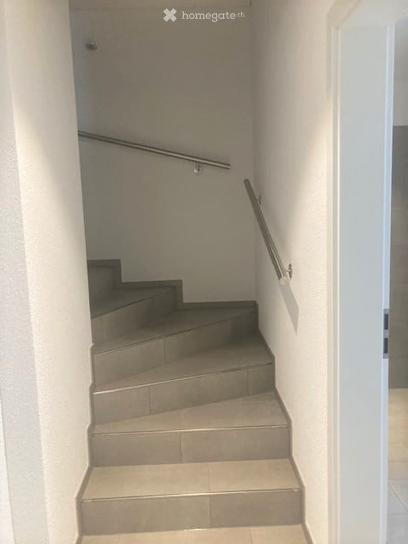 A narrow staircase with concrete steps and metal handrails, leading upwards. The walls are painted white, and the floor is tiled.