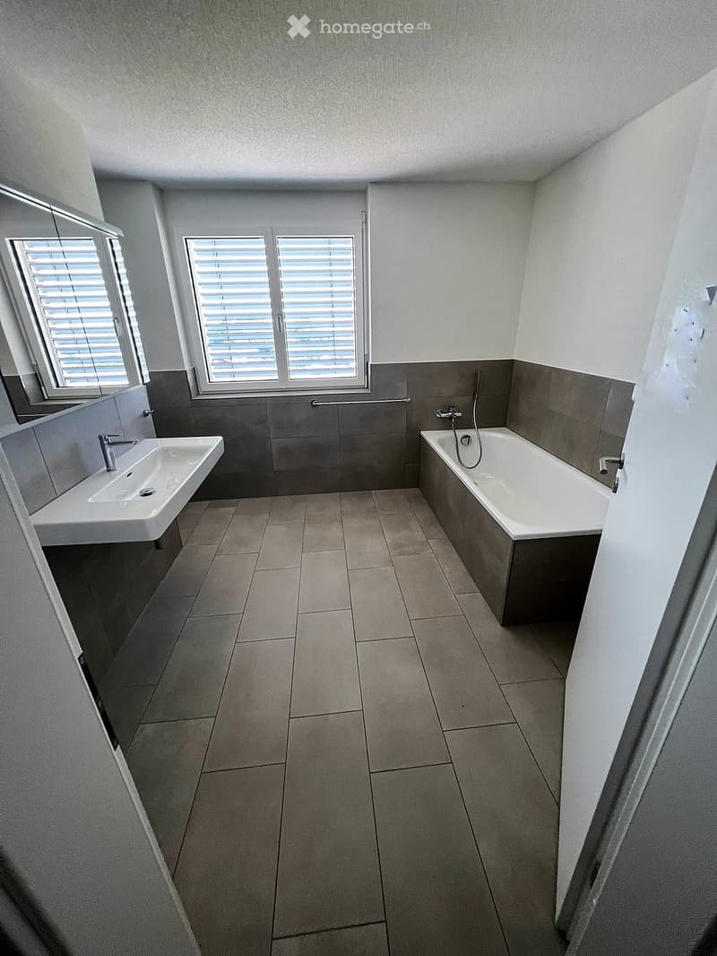 Modern bathroom with a bathtub, sink, and tiled walls. Features large windows with blinds and a clean, minimalist design.