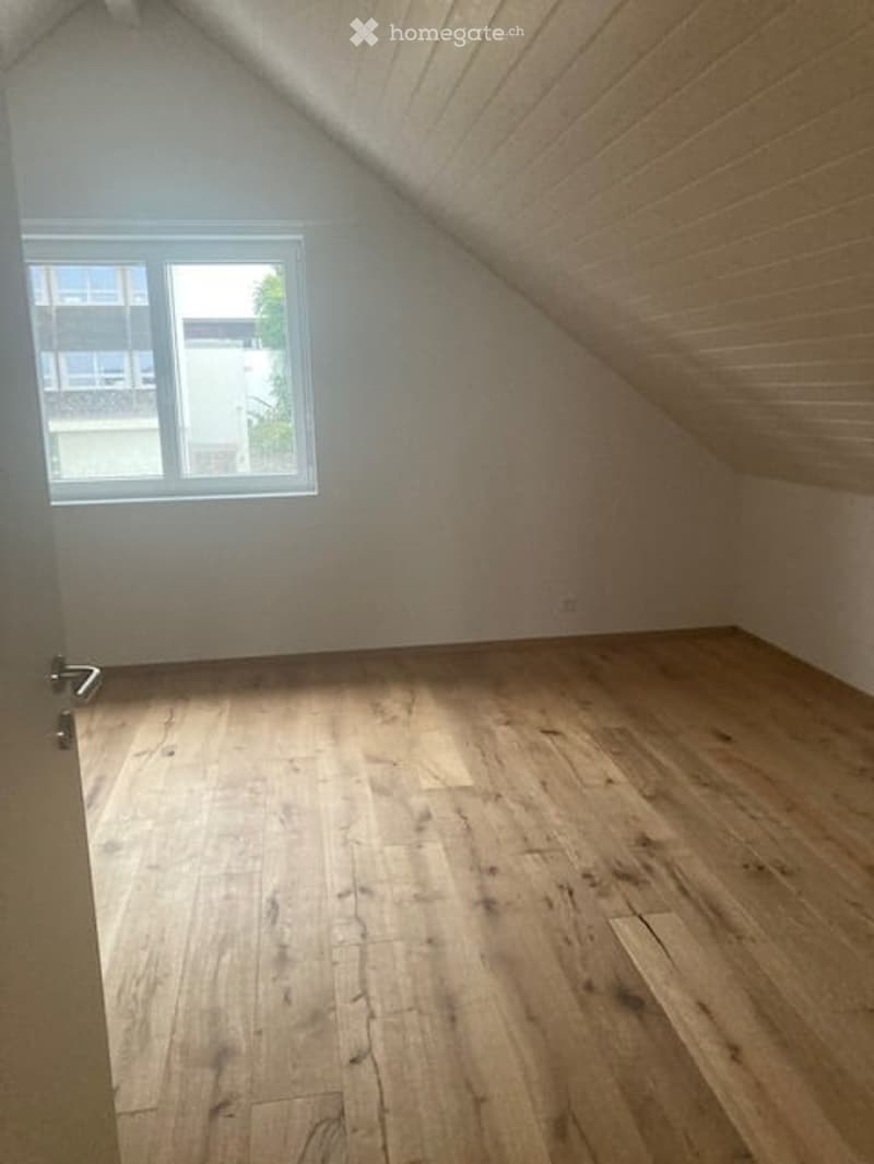 Empty attic room with wooden flooring, white walls, and a single window with white frame. Slanted ceiling with exposed wooden beams.