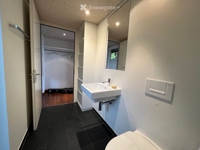 Modern bathroom with white walls, black tiled floor, white sink, mirror, and toilet. An open closet with wooden flooring is visible through the doorway.