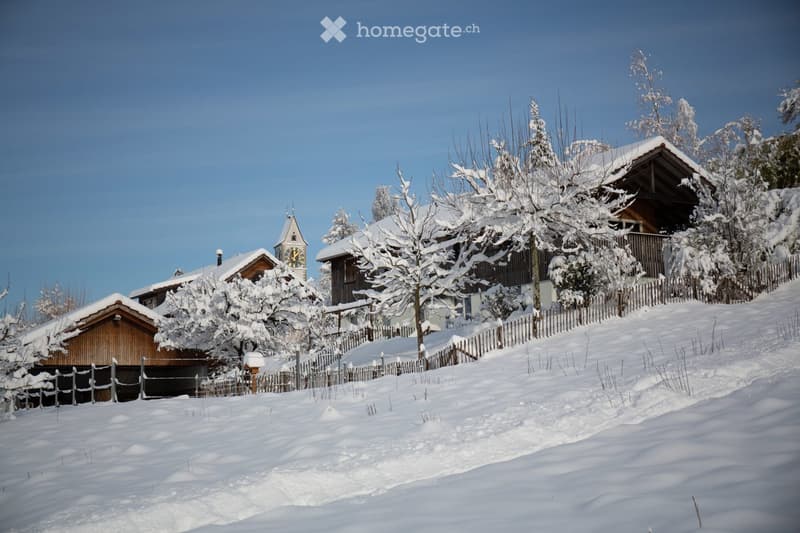 A snow-covered village with traditional wooden houses, a church tower, and a wooden fence lining the pathway.