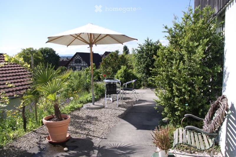 A sunny outdoor patio with a large umbrella, a small table, and chairs, surrounded by greenery and a view of the countryside.