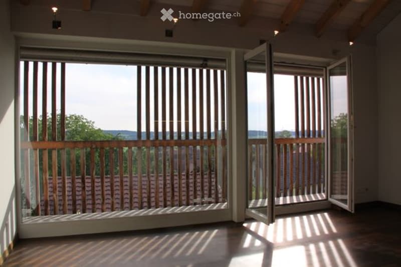 Bright, unfurnished room with large sliding glass doors leading to a balcony, wooden flooring, and exposed wooden beams on the ceiling.