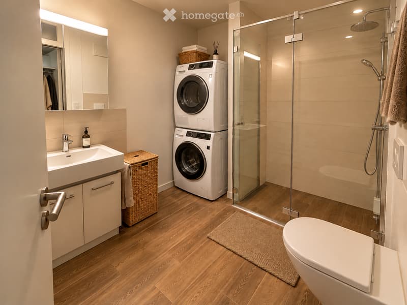 Modern bathroom with a white sink, mirror, toilet, glass-enclosed shower, and stacked washer/dryer unit. Features a wicker basket, towel, and a small rug on the wooden floor.
