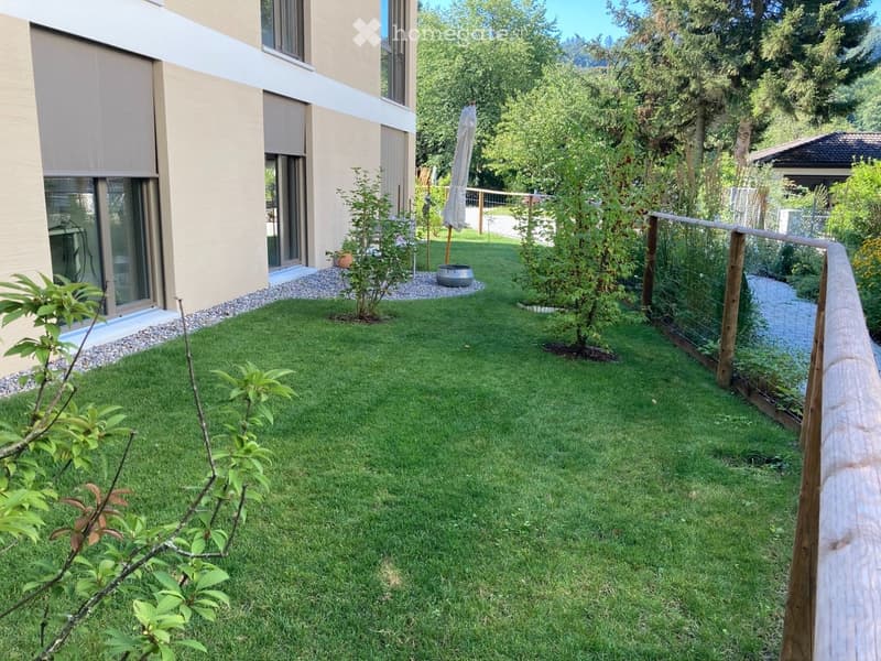Well-maintained garden with green grass, small trees, a wooden fence, and a modern house with large windows and a sliding door.