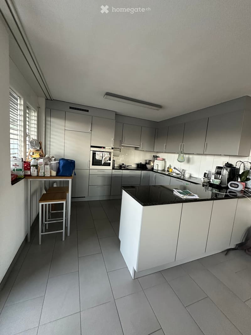 Modern kitchen with grey cabinets, black countertop, and various appliances including an oven, microwave, and coffee maker. A small table with stools is positioned near the window, and various items are placed on the counter and table.