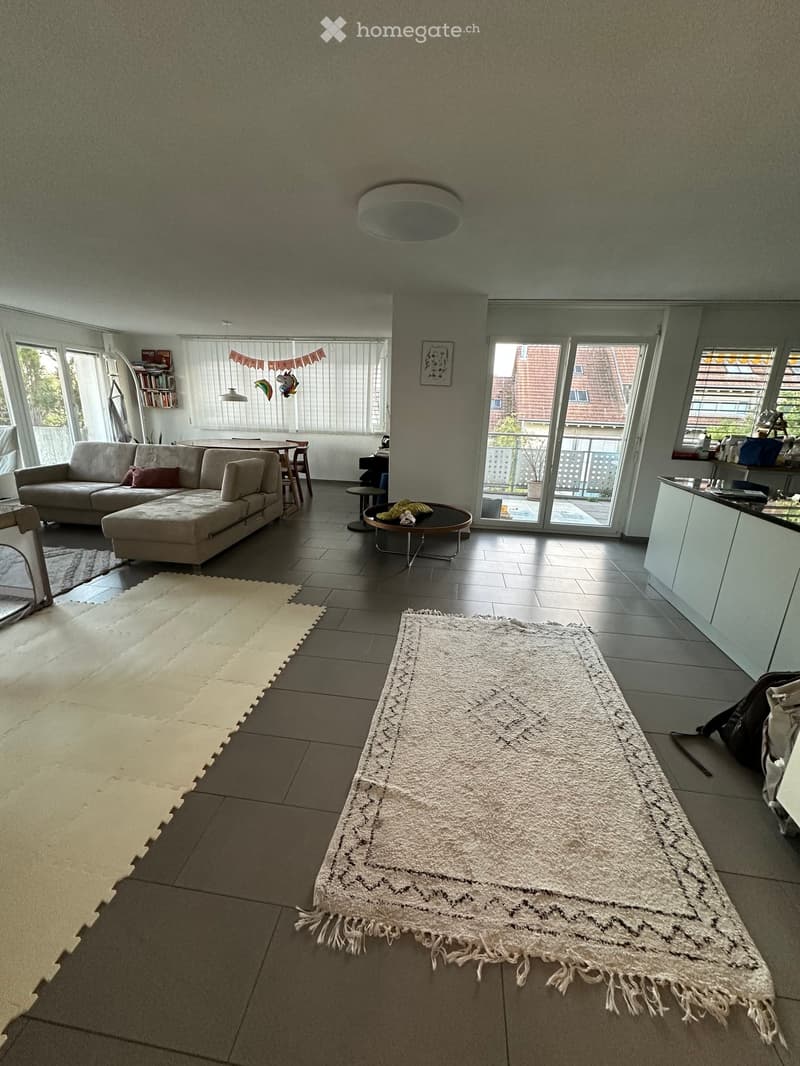 Open-plan living and dining area with a grey sectional sofa, coffee table, dining table, chairs, and a large white rug. Large windows and sliding glass doors lead to a balcony.