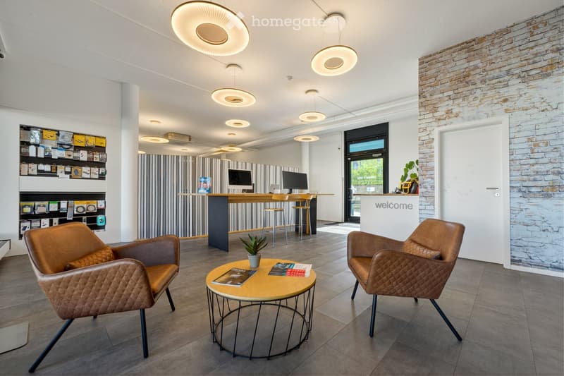 Modern reception area with leather chairs, round coffee table, computer screens, and shelves displaying items.