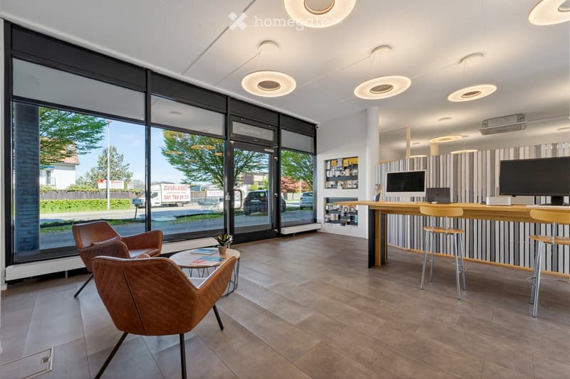 Modern reception area with large windows, leather chairs, a round coffee table, a reception desk with stools, and two computer screens.
