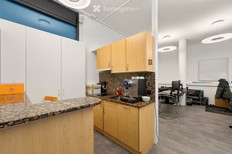The image shows a modern, well-lit kitchen and lounge area. The kitchen features light wooden cabinets, granite countertops, a coffee machine, and a sink. The lounge area includes a sofa, a table with a laptop, and a screen on the wall. The space is brigh