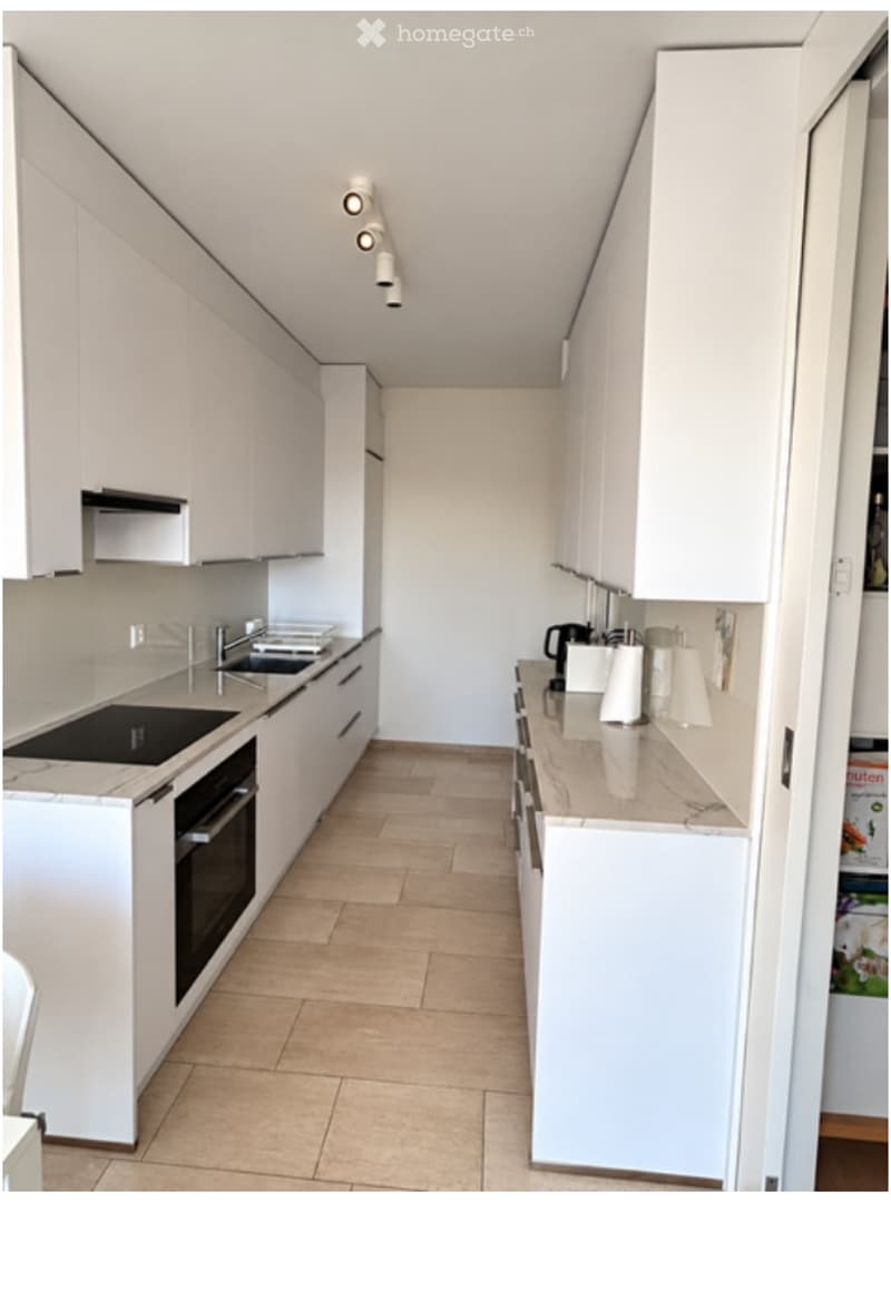 Modern kitchen with white cabinets, a central island, and built-in appliances including a sink, oven, and fridge. The countertops are light-colored, and the floor is tiled.