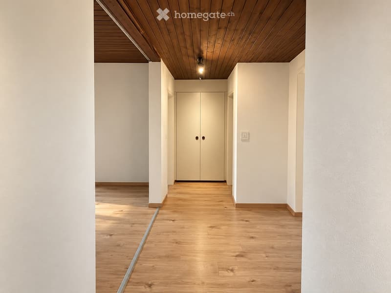 A modern, minimalist hallway with light wooden flooring, white walls, and a double door at the end. The ceiling features wooden paneling and recessed lighting.