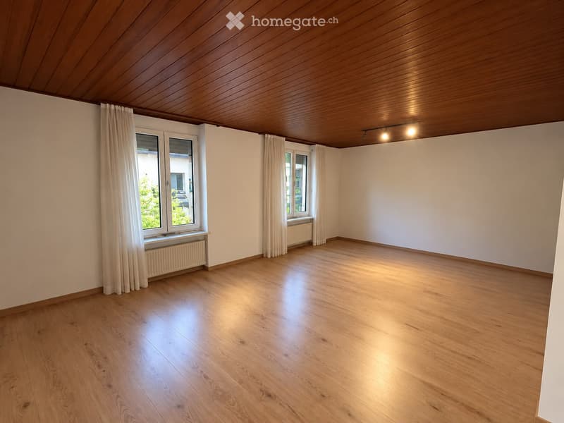 Empty room with wooden flooring, white walls, and large windows with curtains. The ceiling is wooden with recessed lighting.