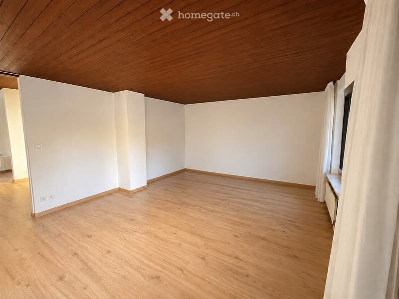 Empty room with light wooden flooring, white walls, and a wooden ceiling. There is a window with white curtains on the right side and an open doorway on the left side.
