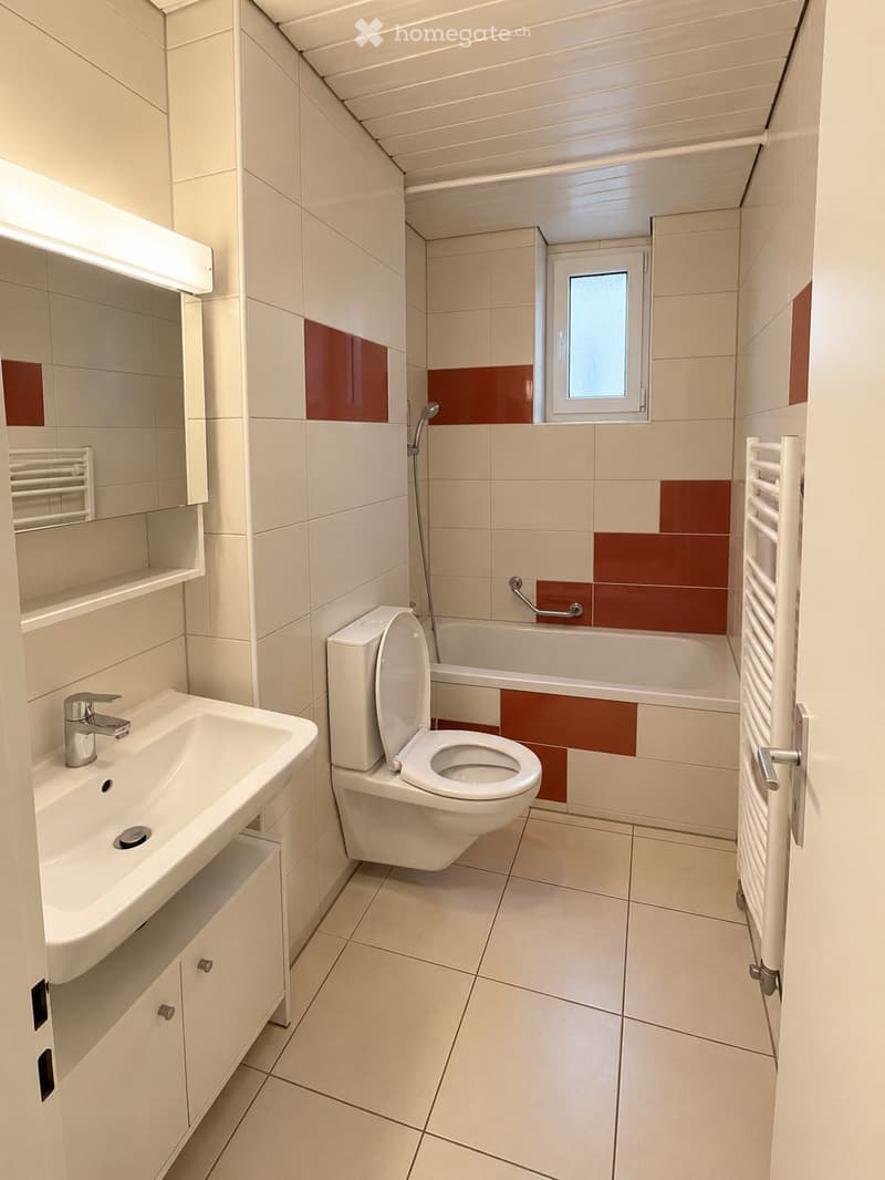 Modern bathroom with white tiles, a sink, toilet, bathtub, mirror, and radiator. Features a window above the bathtub and a built-in shelf with a towel rack.