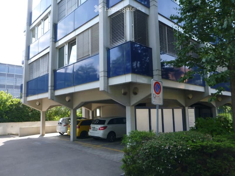 modern building, blue facade, glass windows, covered parking area, yellow and white cars parked, bushes and trees