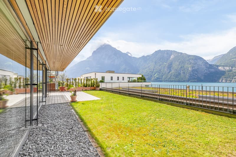 Modern outdoor area with wooden decking, gravel pathway, potted plants, and a view of a lake and mountains.