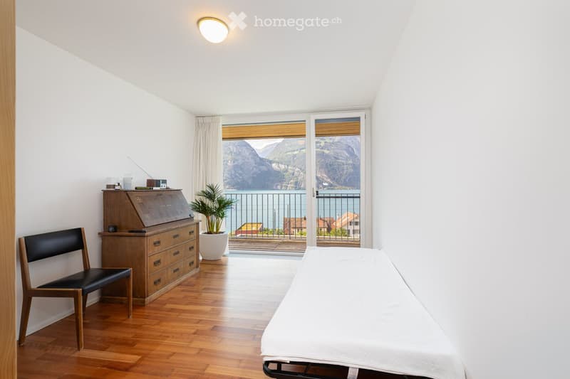 Bright bedroom with wooden flooring, large windows offering a scenic mountain view, a neatly made bed, a wooden desk with a chair, and a potted plant near the window.