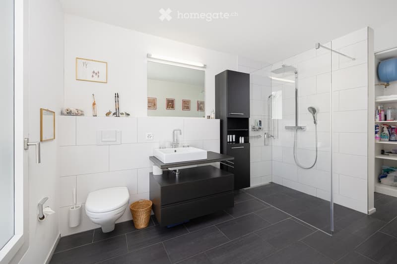 Modern bathroom with white tiles, dark wooden floor, black cabinet, white sink, toilet, shower with glass enclosure, and decorative items on shelves.