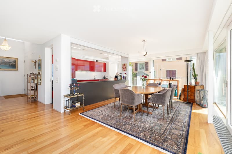 Open-plan living and dining area with wooden flooring, a round dining table with six chairs, a modern kitchen with red cabinets, a floor lamp, and a view through large windows.
