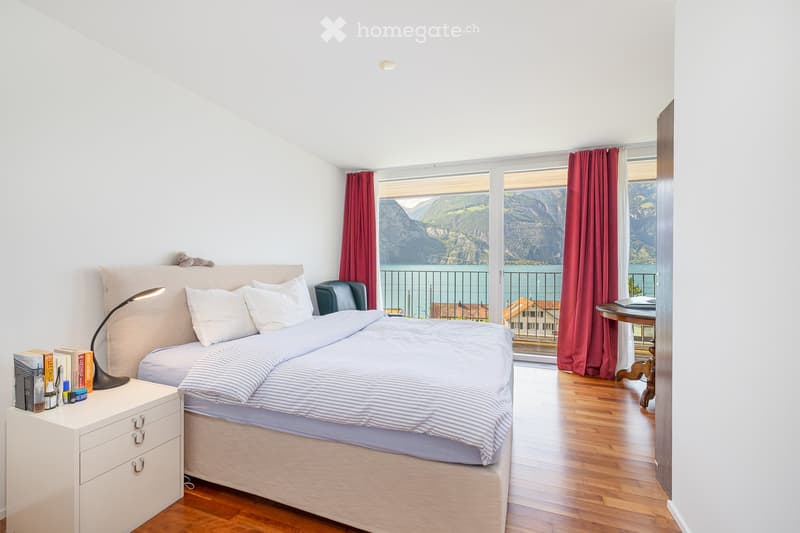 Cozy bedroom with a double bed, nightstand, lamp, chair, and a small table with books. Large windows offer a scenic mountain view.