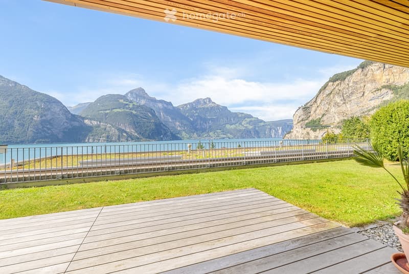 A wooden deck and grassy area overlooking a scenic lake and mountain range, with a metal railing and a covered wooden pergola.