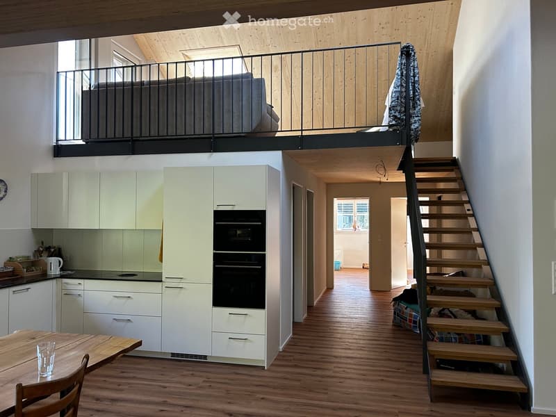 Modern, open-plan living space with a raised sleeping loft, kitchen area, and staircase leading to the loft. Features include a wooden dining table with chairs, white kitchen cabinets, and a black metal balcony railing.