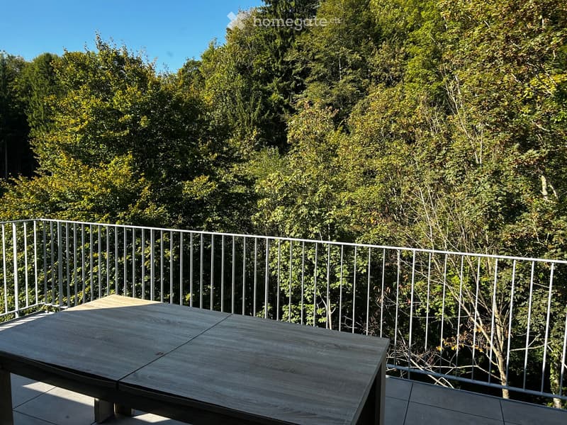 Balcony with a wooden table and a metal railing, surrounded by lush green trees.