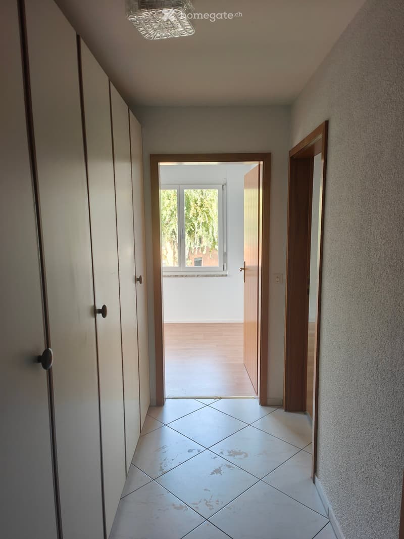 A narrow hallway with tiled flooring, white walls, and wooden doors on both sides, leading to an open area with a window and a view of greenery outside.