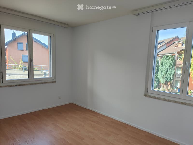 Empty room with light wooden flooring and white walls, featuring large windows with white frames and a view of neighboring buildings and greenery.